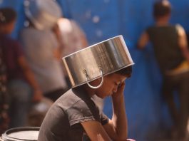 Uma criança colocando uma panela na cabeça para se proteger do sol enquanto espera para pegar comida