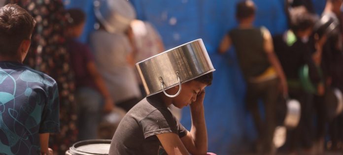 Uma criança colocando uma panela na cabeça para se proteger do sol enquanto espera para pegar comida